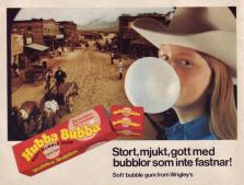 En tjej som tuggar Hubba Bubba och blåser en stor bubbla står framför en scen hämtad från en westernstad med hästar och vagnar